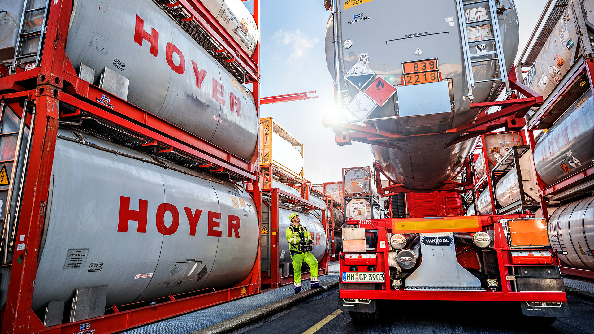 A truck is loaded with a HOYER Group tank container for chemicals. A worker in yellow work clothes stands next to the truck and observes the process.