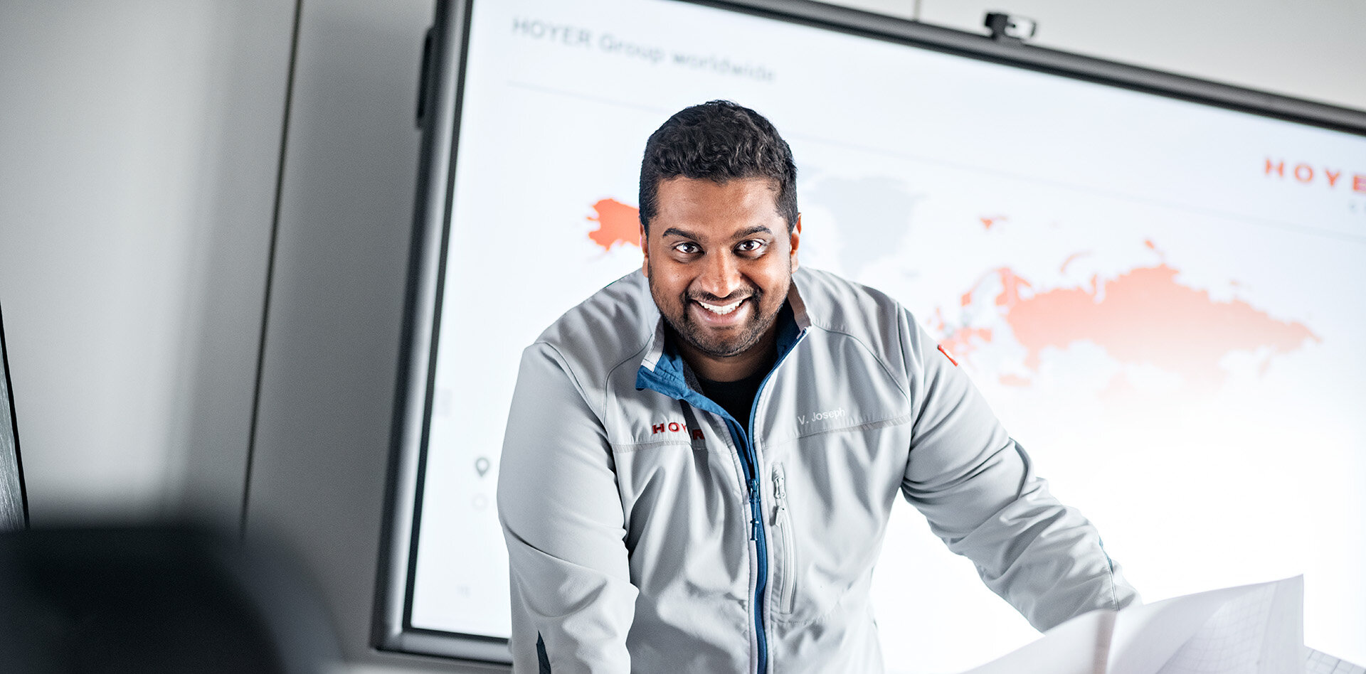 HOYER Mitarbeiter steht in einer HOYER Jacke vor einem Smart Board