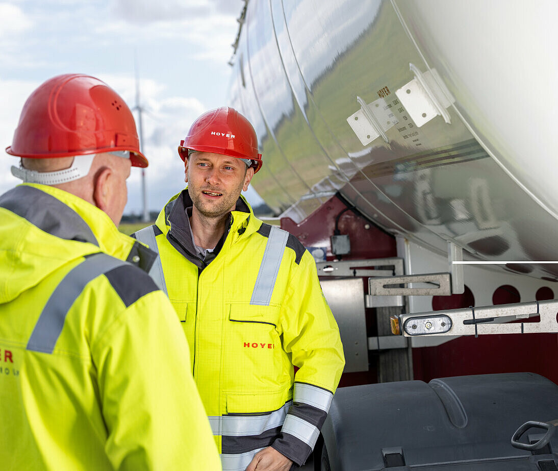 Four HOYER employees in safety clothes squatting in front of a HOYER truck connection and looking at a tablet