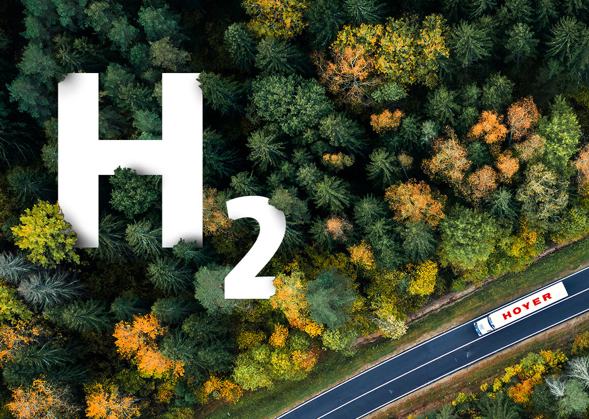 Aerial view of a white HOYER hydrogen transport truck on a road passing a wooded area. A large, white H2 logo pokes out of the forest from above.