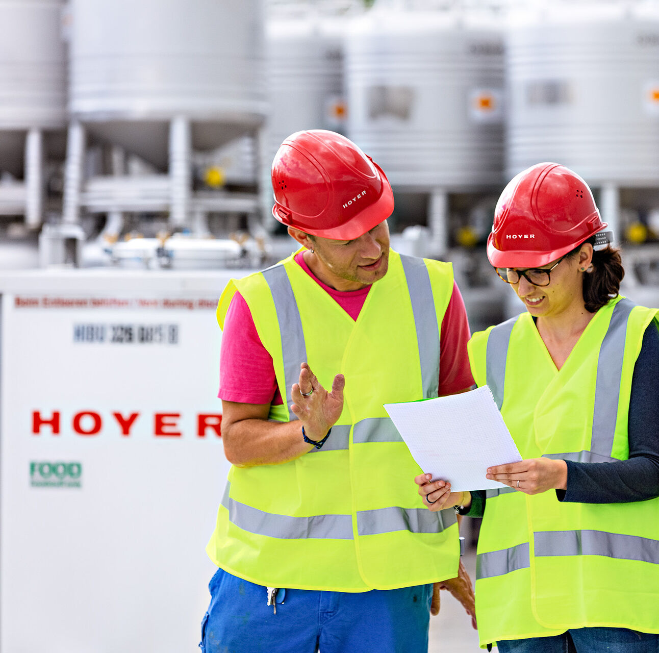 Two HOYER employees in front of a Food IBC and look at a document 