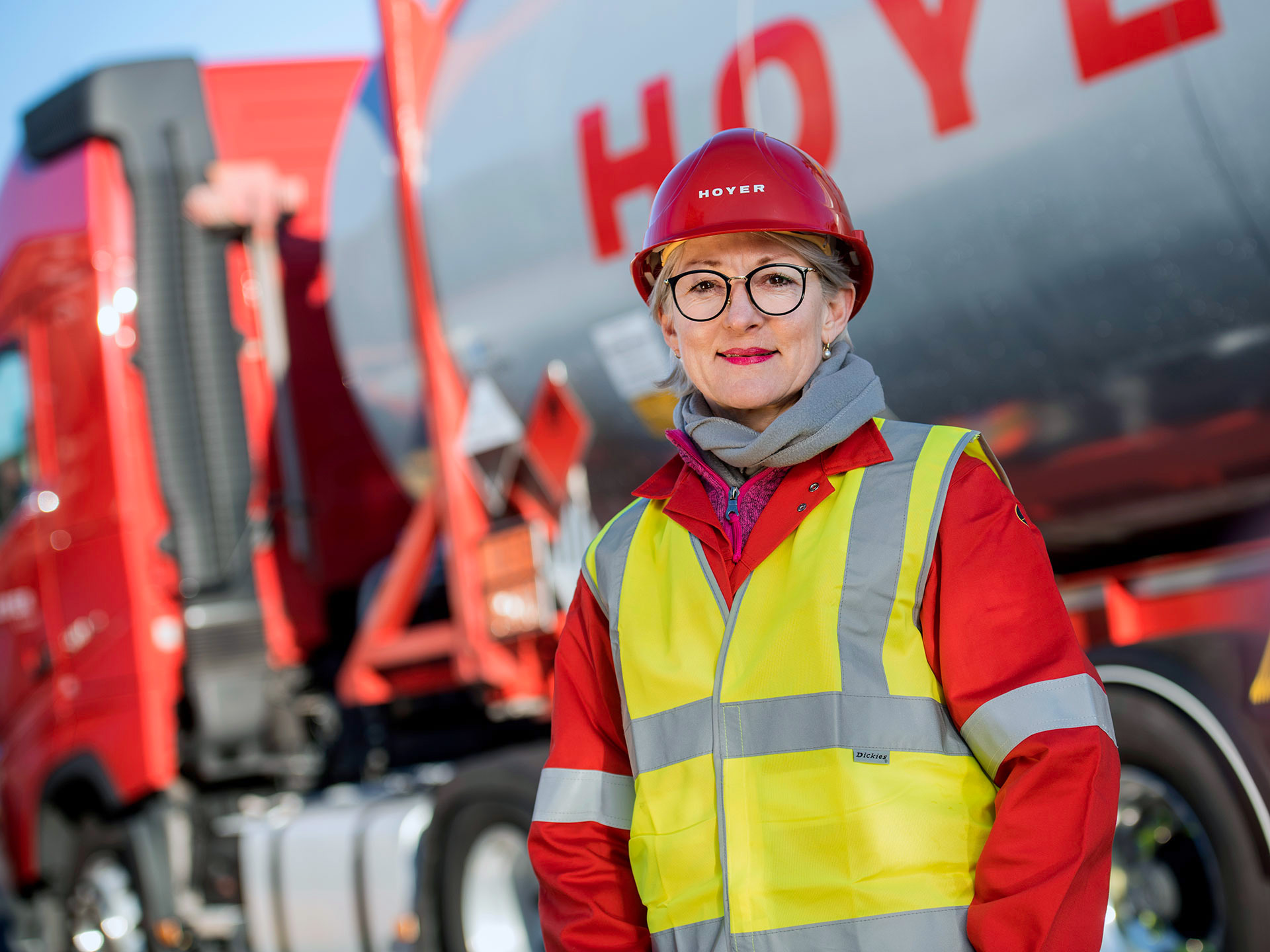 HOYER Mitarbeiterin steht vor einem HOYER Lkw und traegt eine Warnweste und einen roten Helm