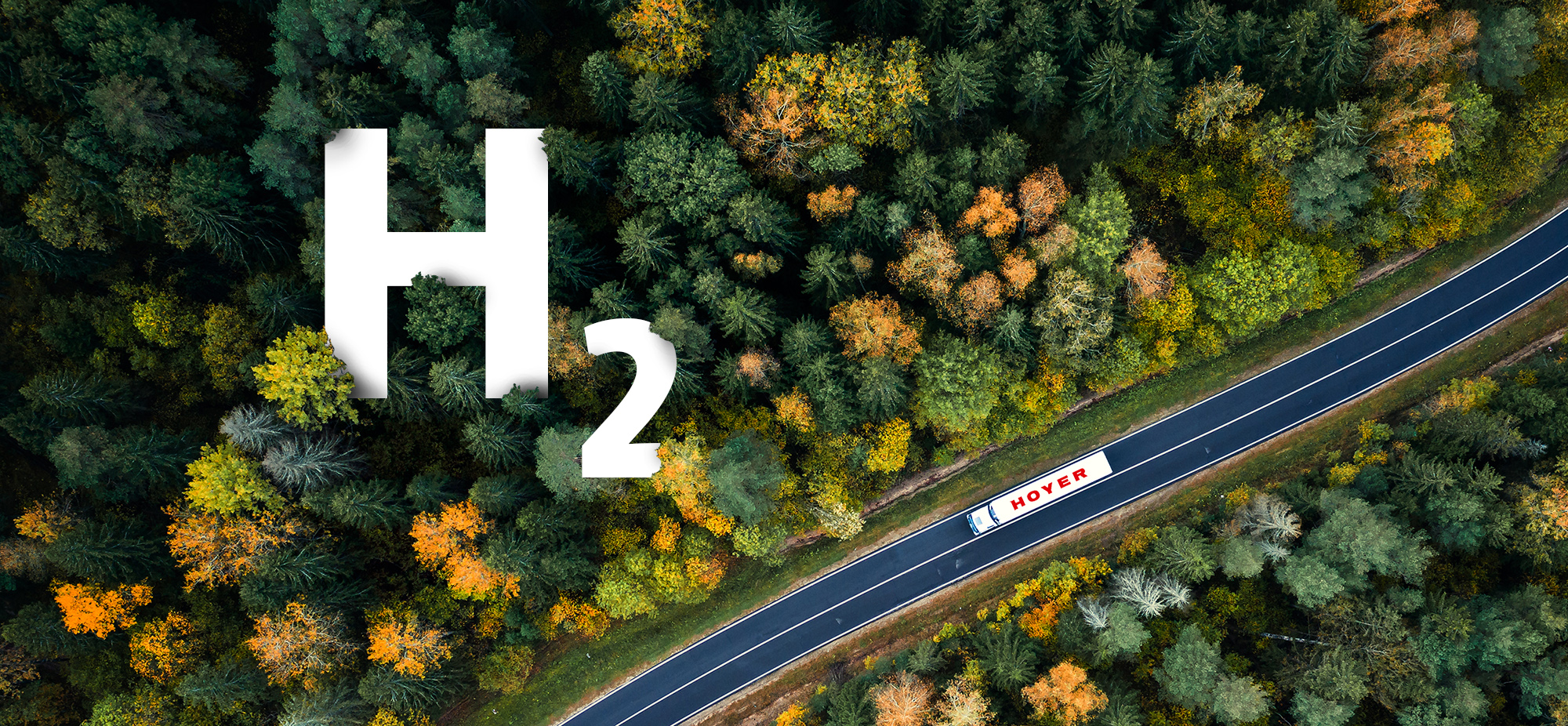 Aerial view of a white HOYER hydrogen transport truck on a road passing a wooded area. A large, white H2 logo pokes out of the forest from above.