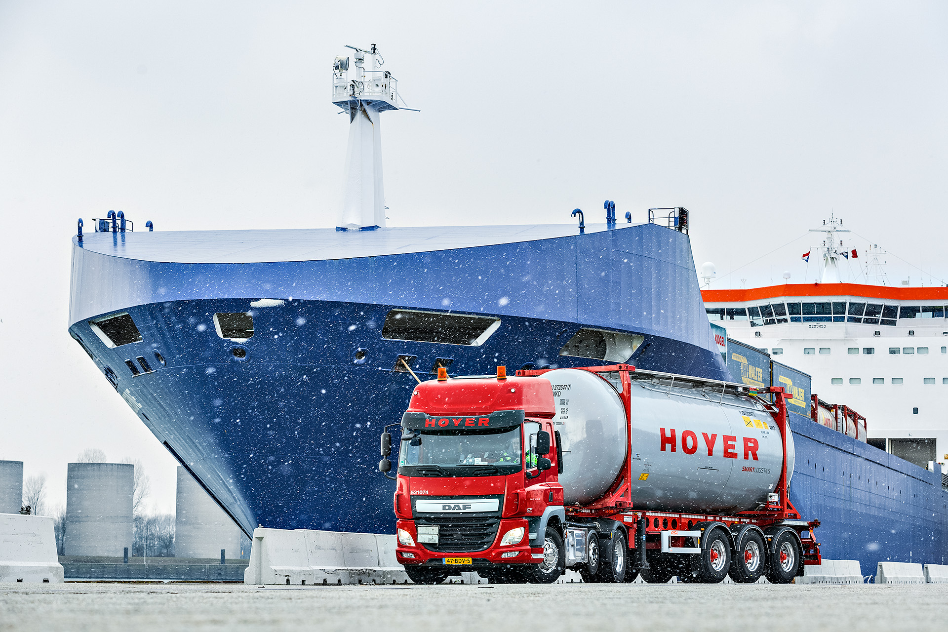 HOYER Lkw mit Chemie-Tankcontainer smart logistics vor einem Schiff. Es schneit.